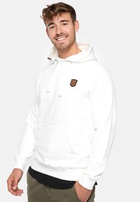 Sudadera blanca con capucha, fabricada en tejido suave, que presenta un bolsillo frontal y un parche de logo marrón. Puños y dobladillo acanalados.
