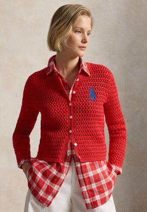 Femme aux cheveux blonds courts portant un cardigan rouge en crochet par-dessus une chemise à carreaux rouge, debout les mains dans les poches, regardant vers la droite.