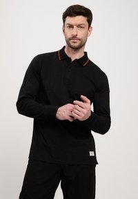 Czarna polo z długim rękawem z pomarańczowymi akcentami kołnierzyka, wykonana z bawełny, z zapinaniem na guziki i małą metką marki na dolnym brzegu.