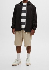 Lätt, svart zip-jacka med huva över en grå och vit randig skjorta. Beige shorts och vita sneakers med grå detaljer kompletterar outfiten.