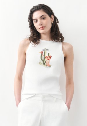 Frau mit lockigem, dunklem Haar, die ein weißes ärmelloses Oberteil mit einem Kaktus- und Stiefelmotiv sowie weiße Hosen trägt und mit den Händen in den Taschen steht.