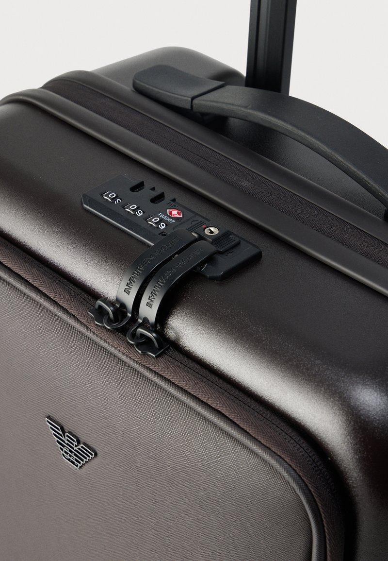 Hard-shell dark brown suitcase with textured finish, combination lock, black zipper pulls, and a retractable handle. Winged logo on front.