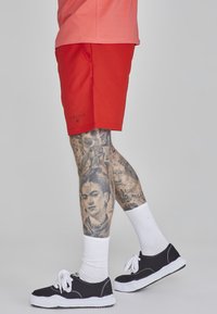 Raudoni maudymosi šortai su šoninėmis kišenėmis ir juodu logo, derinami su juodais sportbačiais ir baltais kojinėmis. Oda turi išsamių tatuiruočių.