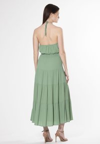 Vestido largo verde con múltiples niveles, escote halter y cintura elástica. La tela es ligera y fluida, realzando su diseño estratificado.