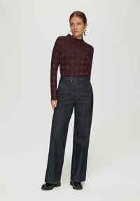 Bordeaux langekleurige top met een houndstooth-patroon, gecombineerd met donkere wijde broek. Model draagt zwarte chunky laarzen en zilveren oorbellen.