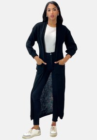 Cardigan largo negro con frente abierto y bolsillos, hecho de un suave material de punto. Lleva puesto sobre una blusa blanca y jeans ajustados negros con zapatillas.