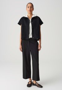 Schwarze ärmellose Jacke mit Kapuze, Knopfverschluss und zwei Fronttaschen, kombiniert mit einem weißen Oberteil, schwarzen weiten Hosen und schwarzen Netzschuhen.