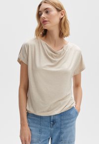 Top beige drappe con maniche corte e texture morbida, caratterizzato da un collo rotondo e una vestibilità ampia, abbinato a jeans blu chiaro.