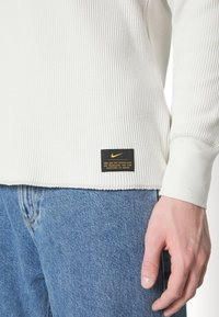 Chemise blanche à manches longues côtelée avec une étiquette Nike noire et orange à l'ourlet. Associée à un jean en denim bleu, mettant en valeur la texture et les coutures.