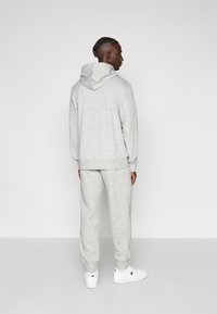 Γκρι φούτερ με τσέπη καγκουρό και ρυθμιζόμενο καπό, συνδυασμένο με αντίστοιχο στενό παντελόνι. Και τα δύο είναι κατασκευασμένα από απαλό, υφασμένο ύφασμα. Λευκά αθλητικά παπούτσια.