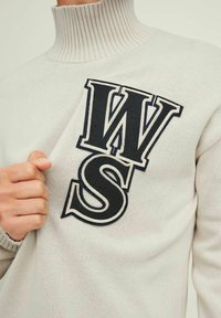 Person som bär en vit ribbad polotröja med stora svarta och vita "WS"-bokstäver på bröstet som drar i tyget något.