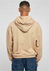 Felpa beige con cappuccio, dal taglio morbido e confortevole, con maniche a costine. Presenta una tasca a marsupio ed è abbinata a jeans blu scuro.