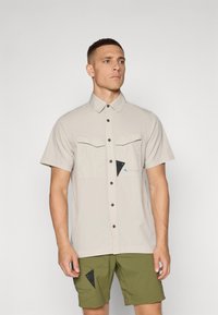 Korte mouw beigekleurig shirt met knoopsluiting, twee borstzakken, zwarte accenten en een blauwe logo detail. Lichtgewicht, gladde stof.