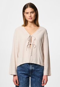 Light pink, textured fabric top with a deep V-neck and tie closure at the front. Features long sleeves and a cropped hem. Worn with blue jeans.