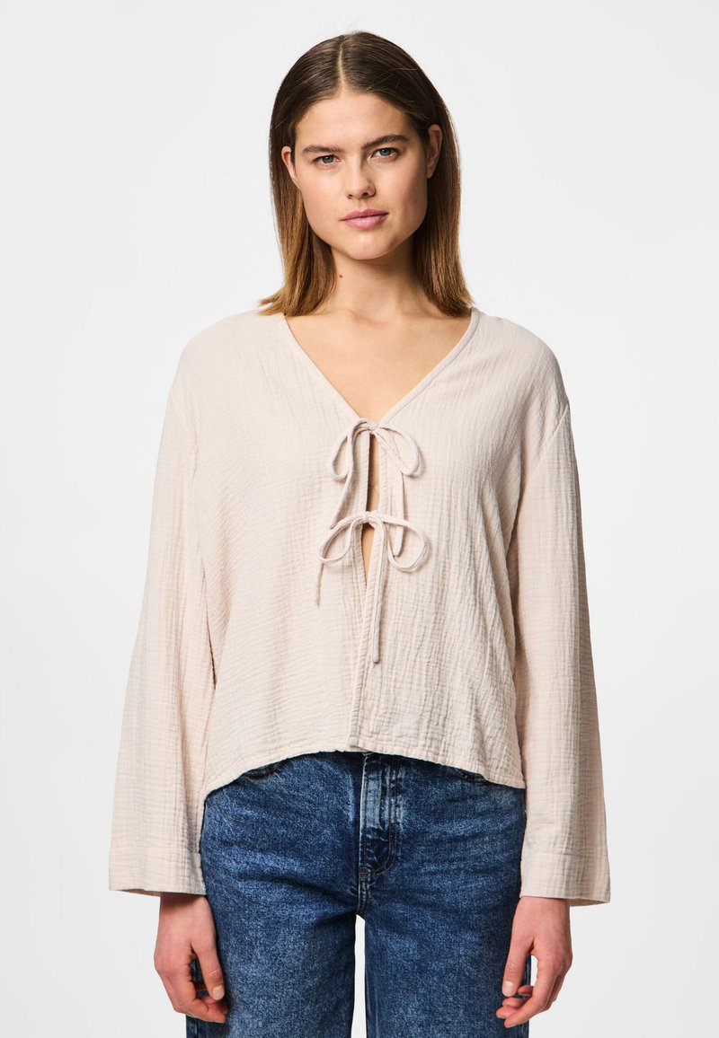 Light pink, textured fabric top with a deep V-neck and tie closure at the front. Features long sleeves and a cropped hem. Worn with blue jeans.