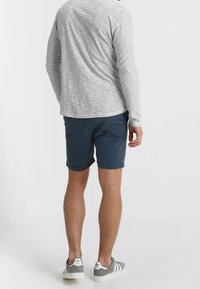 Chemise à manches longues grise à rayures horizontales, associée à un short bleu marine. Les chaussures sont des baskets grises et blanches. Vue de dos.