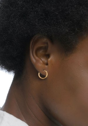 Eine goldene Creole wird aus der Nähe gezeigt, mit einem glatten, runden Design und einem dünnen Profil, die an einem Ohr befestigt ist. Hautfarbe sichtbar.