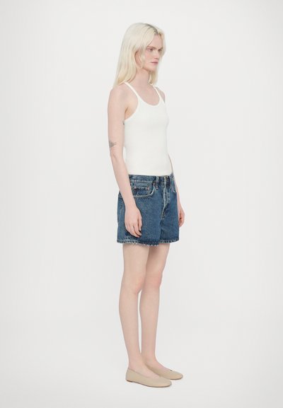 Top sans manches blanc assorti à un short en denim bleu foncé, avec une coupe classique et une finition brute à l'ourlet. Porté avec des ballerines légères.