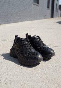 Paire de baskets noires épaisses avec lacets sur un trottoir en béton ensoleillé près d’un mur de bâtiment gris.