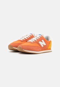 Baskets orange en daim et nylon, avec des lacets blancs, un logo "N" blanc et un accent jaune sur le talon. Semelle légère en caoutchouc.