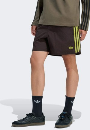 Osoba ubrana w czarne szorty z żółtymi paskami i logo, czarne skarpetki Adidas, czarne buty sportowe oraz oliwkową bluzkę z długim rękawem z ciemnymi paskami.