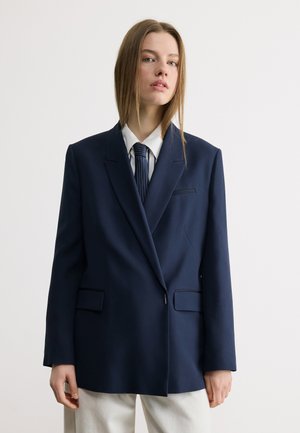 Nuori nainen, jolla on suorat hiukset ja yllään ylisuurikokoinen tummansininen bleiseri, valkoinen paita ja tummanraidallinen solmio vaaleaa, yksinkertaista taustaa vasten.