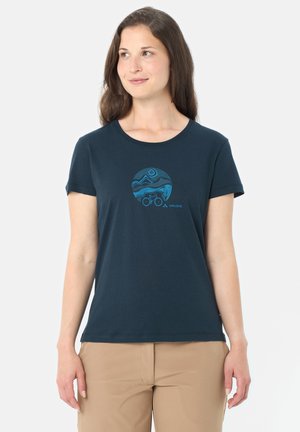 Donna che indossa una t-shirt blu navy con una grafica circolare blu raffigurante una montagna e una bicicletta, abbinata a pantaloni beige, che guarda verso sinistra.