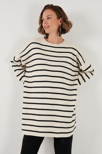 Ženska, oblečena v prevelik bel pulover s črnimi vodoravnimi črtami, črne hlače in črne uhane v obliki cvetja, ki visijo.