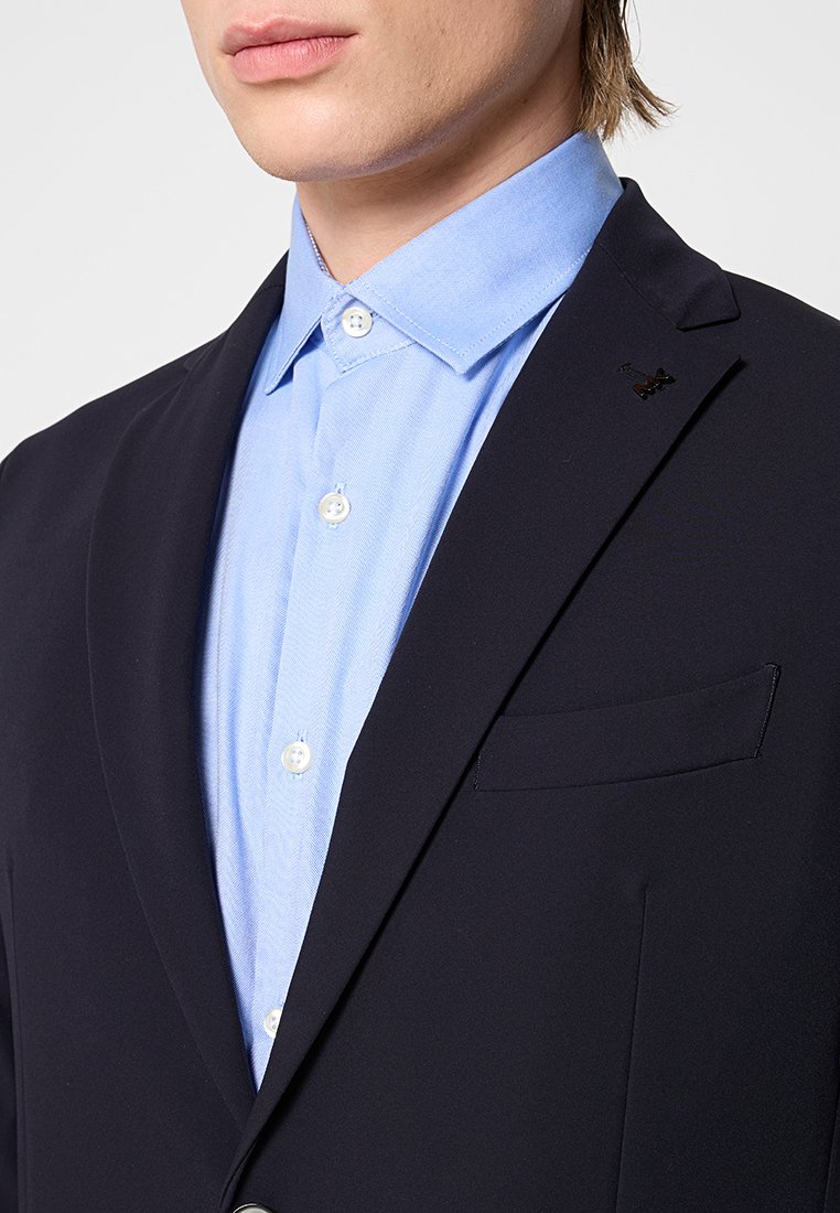 Marineblazer mit taillierter Passform, einer Brusttasche, kombiniert mit einem hellblauen Hemd mit Knöpfen aus strukturierter Baumwolle und weißen Knöpfen.