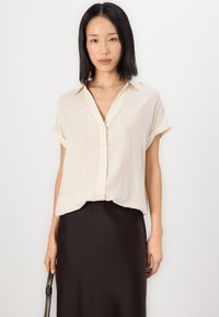 Button-up, short-sleeve blouse in light beige with subtle vertical stripes. Soft fabric, relaxed fit, and a pointed collar. Paired with a black skirt.