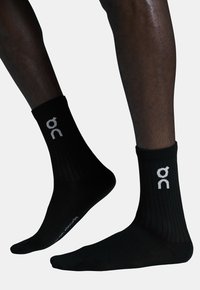 Černé sportovní ponožky s žebrovanou texturou, s bílým logem na lemu a brandingem na podrážce. Design do kotníku.