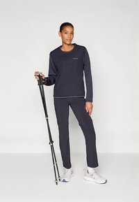 Žena u crnom sportskom odijelu drži crne planinarske štapove, stoji s bijelim tenisicama na jednostavnoj svijetloj pozadini.