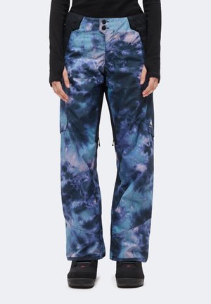 Personne portant un pantalon tie-dye bleu et violet avec des bottes noires, debout devant un fond clair uni.