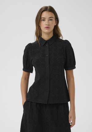 Noor naine kannab musta tekstuuriga nööpidega pluusi koos volangilise lühikese võrguga ja sobivat seelikut heleda tausta ees.