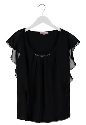 Frau trägt eine schwarze Bluse mit Schmetterlingsärmeln, weiße schmale Hose, schwarze Sandaletten mit Absatz und hält eine schwarze Handtasche.