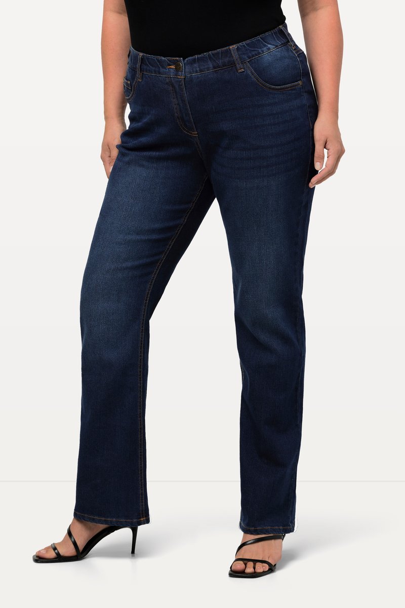 Personne portant un jean bootcut bleu foncé et des sandales à talons noires à brides, debout devant un fond clair uni.