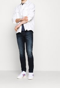 Vit lättviktig skjorta, marinblå t-shirt, mörka jeans med en blekt textur och vita sneakers med röda och blå ränder.