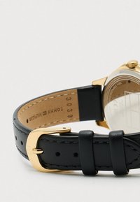 Sort læderurrem med guldhardware; har en glat overflade, en tekstureret underside og en brun inderside.