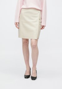 Osoba stojící před jednobarevným pozadím, oblečená v béžové sukni po kolena, růžovém svetru s dlouhým rukávem a černých botách na vysokém podpatku.