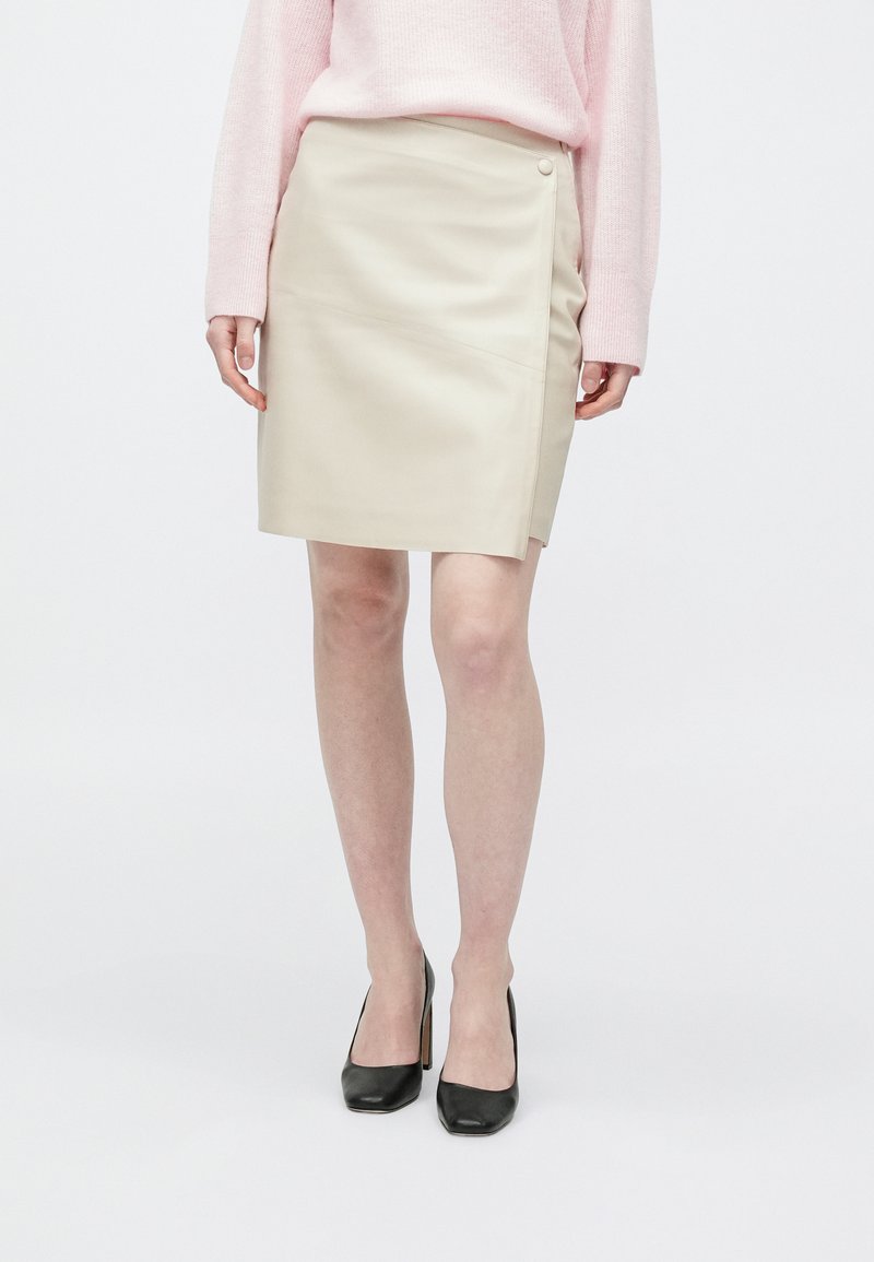 Osoba stojící před jednobarevným pozadím, oblečená v béžové sukni po kolena, růžovém svetru s dlouhým rukávem a černých botách na vysokém podpatku.