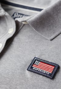 Grå polo skjorte med tekstureret stof, der har et marineblåt badge med et broderet amerikansk flag og teksten "SUPERDRY" nedenfor. Tre knapper synlige.