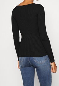 Schwarzes ribbeltes Langarmoberteil mit tailliertem Design und weit ausgeschnittenem Halsausschnitt. Getragen mit blauen Jeans, die eine mittlere Taillenhöhe zeigen.