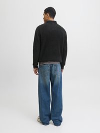 Mann steht mit dem Rücken zur Kamera, trägt einen schwarzen Pullover, blaue Jeans mit weitem Bein, kariertes Hemd und weiße Turnschuhe vor weißem Hintergrund.