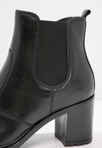 Bottine noire en cuir avec panneau latéral élastique, languette à l'arrière et talon bloc épais sur fond blanc.