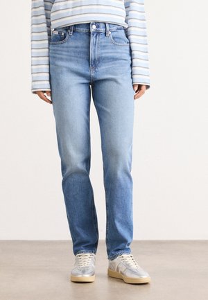 Person iført lyseblå jeans med lige ben, hvide sneakers og en langærmet blå og hvid stribet skjorte, der står på et lyst gulv.