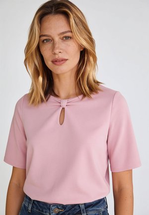 Femme aux cheveux blonds mi-longs porte un haut rose clair à manches courtes avec un petit trou en forme de serrure à l’avant et un détail nœud.