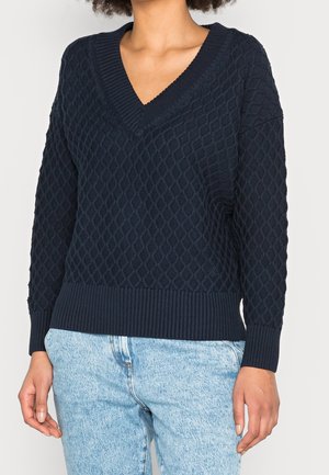 Mørkeblå strikket sweater med diamantmønster, V-udskæring, ribkant ved nederste kant og ærmer. Stoffet ser tekstureret og varmt ud.