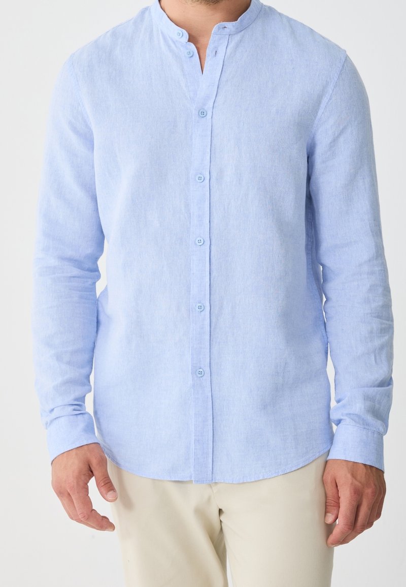 Homme portant une chemise bleu clair à manches longues avec col mao boutonnée et un pantalon beige, debout les bras détendus le long du corps.