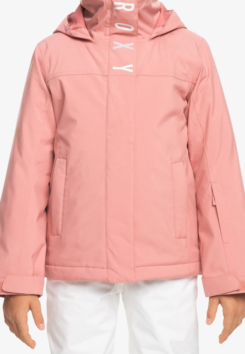 Veste à capuche rose avec fermeture éclair et rabat, poches avant et sangles réglables aux poignets, portée par-dessus un pantalon blanc.