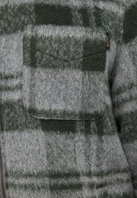 Graues und grünes kariertes Hemd mit flauschiger Textur, mit einer Brusttasche und einem sichtbaren Reißverschluss. Das Etikett trägt die Aufschrift "MUSTARD".